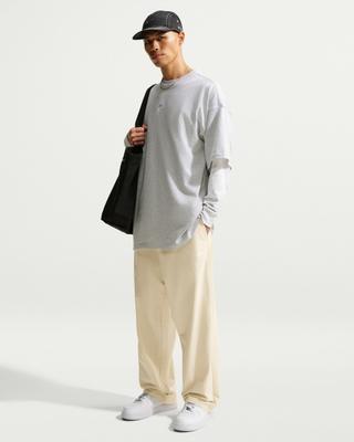 A man wearing a black Nike five-panel cap, a grey oversized Nike tee layered over a light grey long-sleeve top, cream straight-leg Club pants and white Air Force 1 sneakers, carrying a black tote bag over his shoulder against a white studio backdrop.