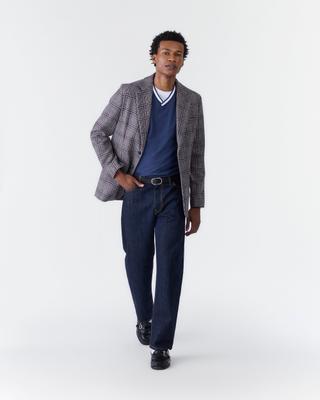 A man wearing a grey-and-burgundy check sport coat over a navy V-neck sweater with white tipping and a white tee, dark raw-denim straight jeans with a black belt, and black loafers, walking toward the camera with one hand in his pocket against a pale grey studio backdrop.