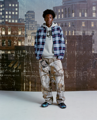 A man wearing a blue-and-red plaid flannel jacket over a grey graphic hoodie, baggy light Realtree-camo jeans and iridescent blue-purple sneakers, holding a black skeleton-hand wallet at his hip, standing full length before a painted, weathered New York skyline backdrop.