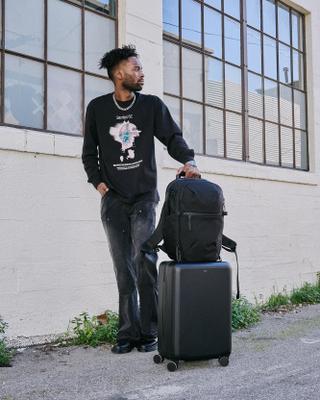 A man wearing a black graphic long-sleeve tee, faded black carpenter jeans and black leather shoes rests his hand on a black backpack set on top of a black hardshell carry-on, standing against a white-painted brick wall.