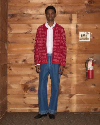 A man wearing a red jacquard cardigan with cream rodeo-themed text over a white button-up shirt, mid-blue flared jeans with decorative stitched hems, and black loafers, standing full length against a wood-panelled barn wall.
