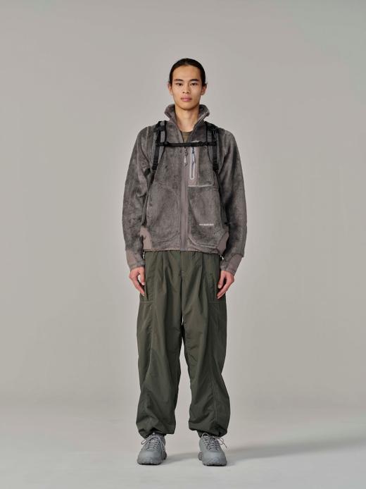 A man wearing a grey high-pile fleece zip jacket over an olive base layer, with a black-strapped backpack, loose olive cargo trousers and grey trail shoes.