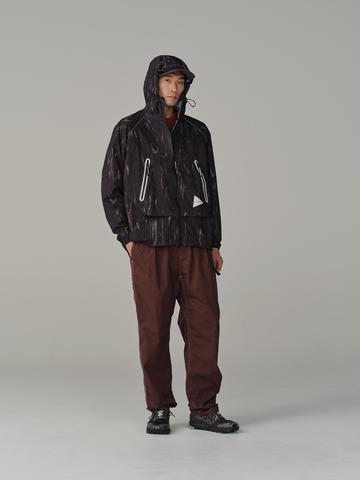 A man wearing a dark printed hooded wind jacket with zip chest pockets, wide burgundy cotton trousers, and black hiking shoes.