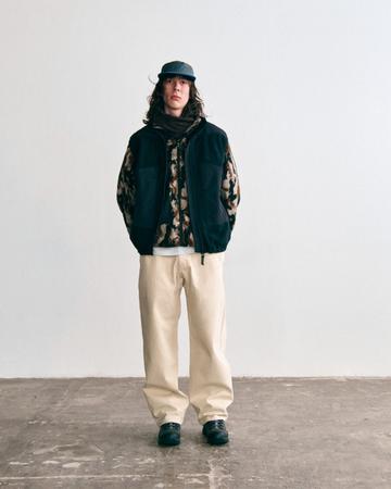 A man wearing a camo-print fleece jacket under a black fleece vest, a dark fleece neck scarf, a nylon cap with blue trim, cream twill pants and black low hiking shoes, standing in front of a white wall on a concrete floor.
