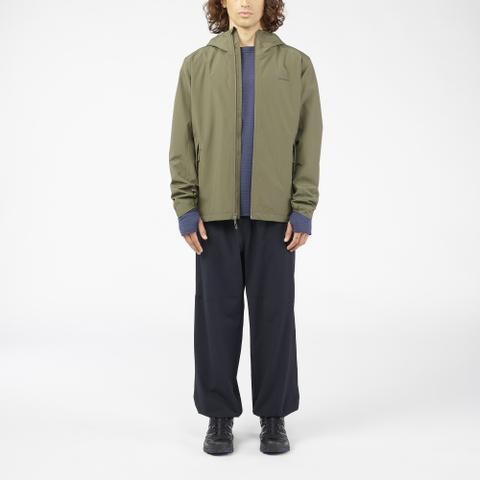 A man wearing an olive hooded shell jacket open over a navy knit midlayer, wide navy trousers and black lugged boots