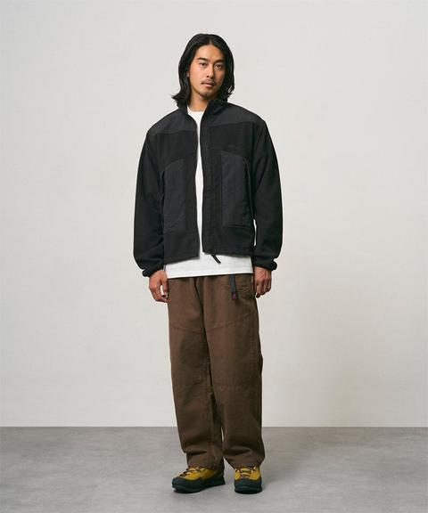A man wearing a black fleece jacket open over a white tee with brown webbing-belt climbing pants worn full-length and yellow low hiking shoes