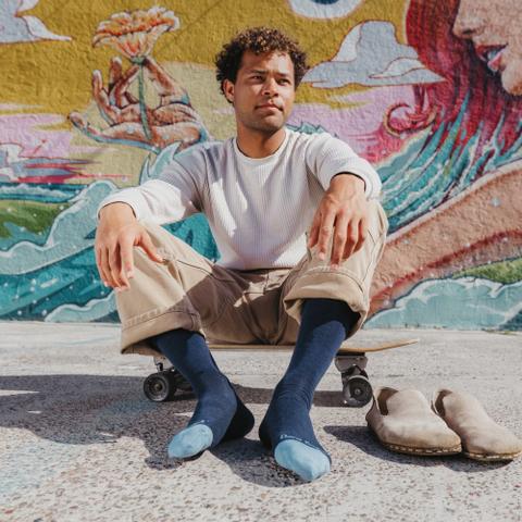 A man wearing a ribbed cream long-sleeve tee and tan cargo trousers, seated on a skateboard in navy crew socks with suede clogs set on the pavement beside him