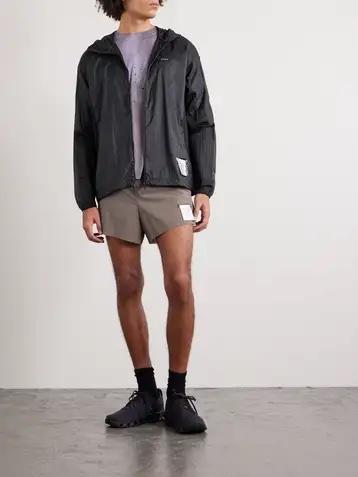 A man wearing a black hooded wind jacket open over a grey technical tee, taupe running shorts, black crew socks and black trail running shoes
