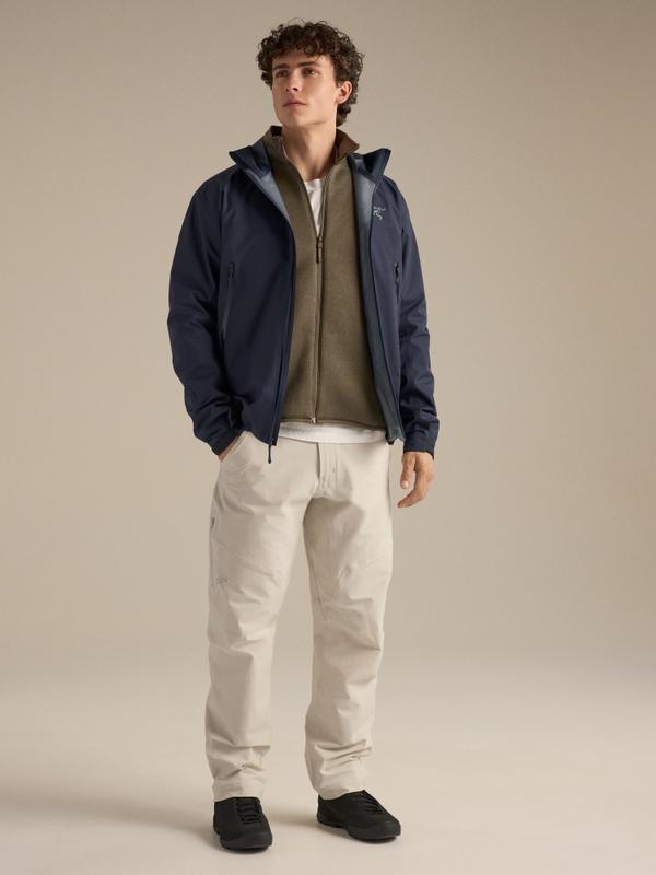 A man wearing a navy Arc'teryx shell jacket open over an olive zip fleece and white T-shirt, with stone-coloured technical trousers and dark trail shoes, standing in a studio.