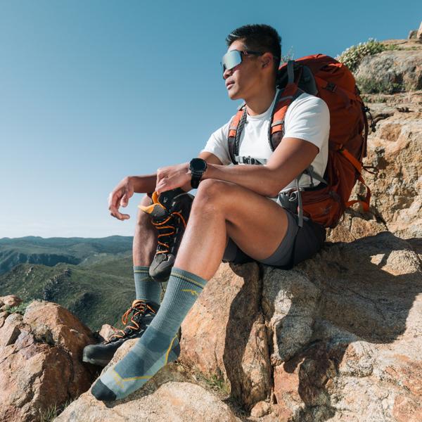 A man wearing a white tee, gray shorts, an orange hiking pack and seafoam boot-height hiking socks sits on a sunlit summit rock, one foot in a trail shoe and the other shoe in his hand, with green mountains behind him.