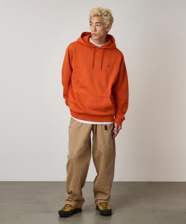 A man wearing a burnt-orange pullover hoodie over a white tee, relaxed tan Gramicci pants and yellow-and-black approach shoes, standing full length in a studio.