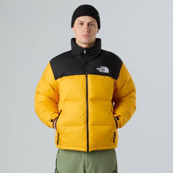 A man wearing a Summit Gold and black 1996 Retro Nuptse down jacket zipped up, with a black beanie and green trousers, hands in pockets, against a light grey studio background.