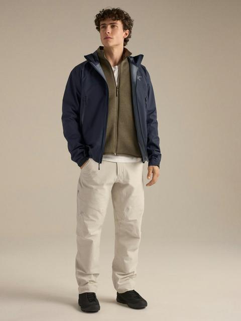 A man wearing a navy hooded hard shell jacket open over an olive zip fleece, cream technical trousers and low black approach shoes