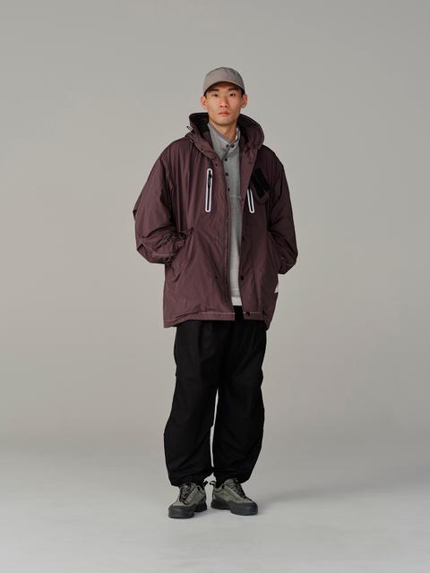 A man wearing a plum hooded technical shell open over a grey half-button pullover, wide black cuffed nylon trousers, grey-green suede trail shoes and a grey cap, standing with hands in pockets against a plain grey studio backdrop.