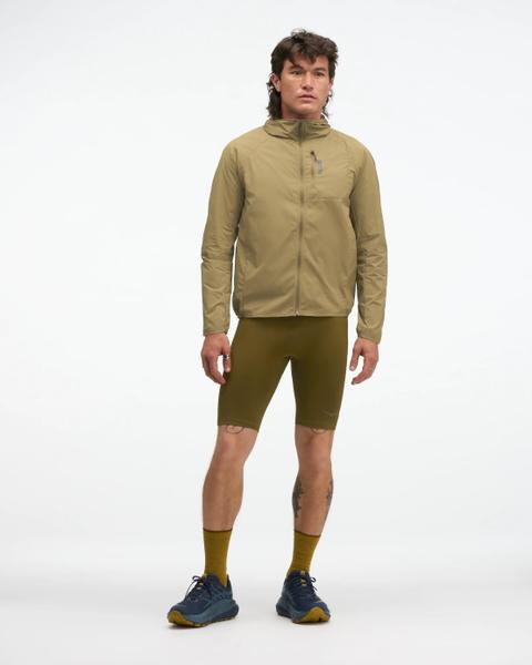 A man wearing a tan hooded wind jacket with olive compression short tights, mustard crew socks and navy trail running shoes, full body, studio backdrop