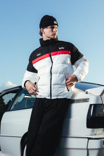 A man wearing a zipped white, black and red Huffer x Audi puffer jacket, a black beanie and wide black trousers, leaning against a white Audi under a clear evening sky.