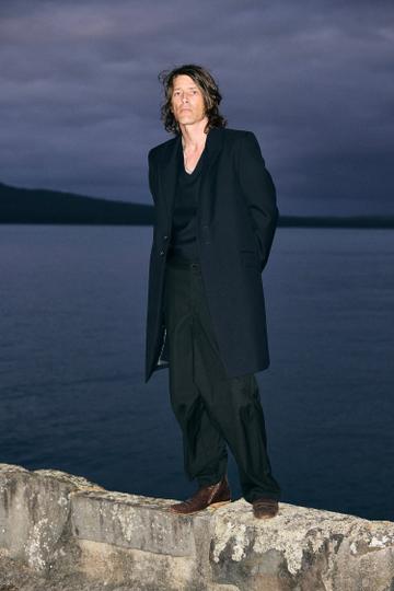 A man with shoulder-length hair wearing a long black tailored coat over a black V-neck top, wide black trousers and brown leather boots, standing on a lichen-covered stone sea wall at dusk with dark water and hills behind him.