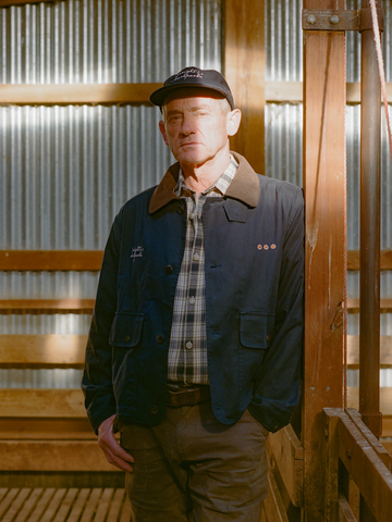 A man wearing a navy waxed-canvas hunting coat with a brown cord collar over a plaid flannel shirt and a black six-panel cap, standing in a sunlit corrugated-iron woolshed.