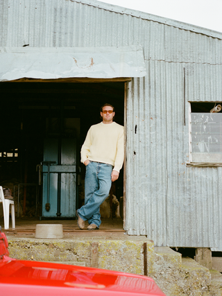 A man wearing a butter-cream crewneck sweater, light-wash straight jeans and tan suede boots, with sunglasses, leaning against the doorframe of a corrugated-iron woolshed.