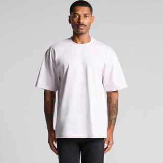 A man wearing a plain pale-lilac boxy heavyweight t-shirt with dropped shoulders and black trousers, standing against a grey studio background