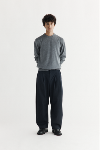 A man wearing a grey merino knitted crewneck on its own with navy double-pleated wide-leg trousers and black loafers, standing full length against a white studio background.