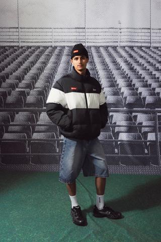A man wearing a zipped black-and-white Huffer x Steinlager Rider puffer jacket, a black beanie, baggy light-wash denim jorts, white crew socks and black runners, standing full length with his hands in his pockets on green turf in front of a black-and-white stadium-seating backdrop.