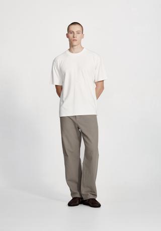 A man wearing a plain off-white short-sleeve tee, straight-leg grey work pants and dark brown low shoes, standing full length with his hands behind his back against a white studio background.