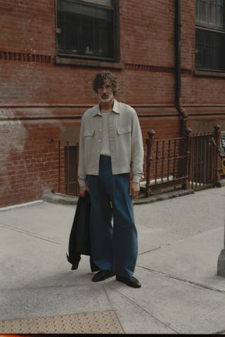 A man wearing a stone blouson shirt over a cream knit vest, wide blue pleated trousers and black loafers, holding a black jacket on a city pavement beside a brick building.