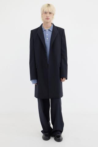A man with platinum blond hair wearing a navy-black knee-length tailored coat over a pale blue ribbed button polo, with wide-leg dark trousers and black leather shoes, standing full length against a white studio background.