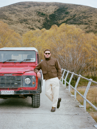 A man wearing a brown cropped hunting coat over a dark roll-neck, cream trousers, lace-up hiking boots and sunglasses, leaning on a red Land Rover Defender on a bridge with autumn willows and a tussock hill behind.