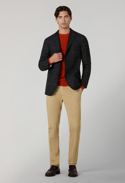 A man wearing an unbuttoned charcoal check wool jacket over a rust-orange crewneck knit, slim tan cotton trousers, dark socks and dark burgundy polished-leather penny loafers, standing against a plain light-grey background.