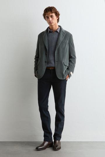 A man wearing a smoky-grey corduroy unstructured jacket open over a grey crewneck knit and a small-check collared shirt, with navy flat-front cotton trousers, a brown leather belt, and dark brown polished leather chelsea boots, standing against a pale studio wall.