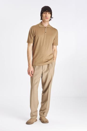 A man wearing a camel fine-knit short-sleeve polo, pleated beige wool trousers with turn-ups, worn-in tan suede slip-on shoes and a dark brown knit beanie, standing on a plain white studio background.