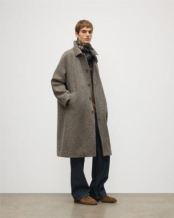 A man wearing a relaxed grey-brown herringbone tweed coat buttoned through the chest and open below, over a brown knit and a plaid wool scarf, with wide dark-indigo jeans and tan suede moc-toe shoes, standing in a white studio with a concrete floor.
