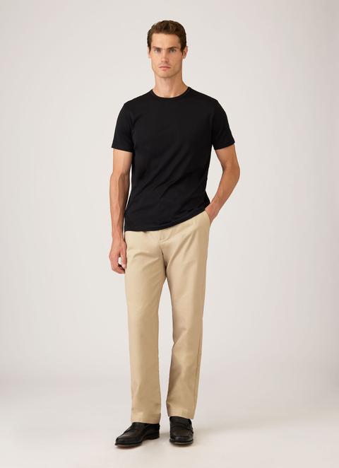 A man wearing a plain black crew-neck T-shirt, light stone cotton chinos and black leather penny loafers, standing with one hand in his pocket against a plain light-grey background.