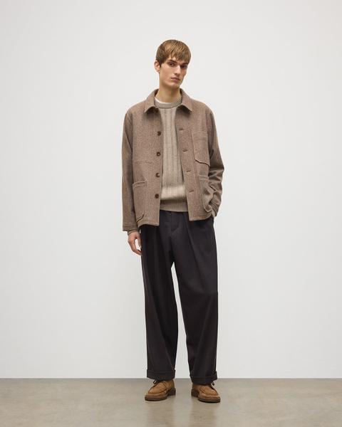 A man wearing an open camel-brown textured wool chore jacket over an oatmeal ribbed crewneck sweater, wide dark-brown pleated wool trousers with turn-ups, and tan suede moc-toe lace-up shoes, standing on a white-and-concrete studio set.
