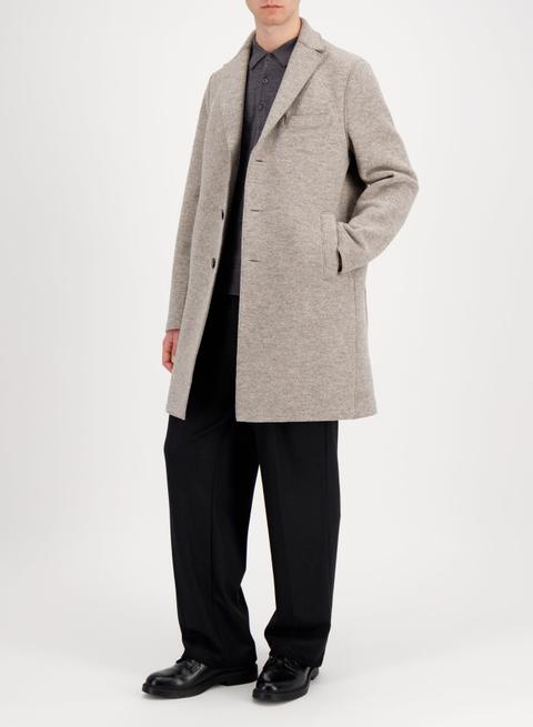 A man wearing an oatmeal-grey pressed-wool single-breasted overcoat worn unbuttoned over a charcoal fine-knit polo, with wide black pleated trousers and black polished leather derby shoes.