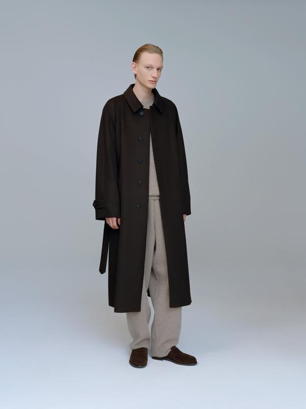 A man wearing a long dark brown wool coat, unbelted with the belt hanging at his side, over an oatmeal knit top and matching wide trousers, with brown suede loafers, standing against a plain grey studio backdrop.