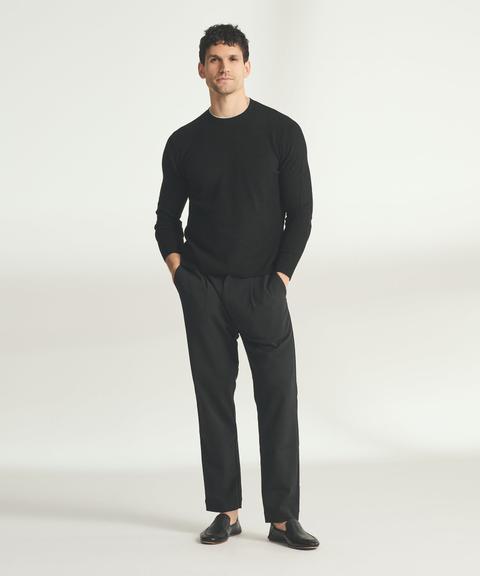 A man wearing a black lightweight cashmere crewneck with thin cream tipping at the collar, black pleated tapered trousers and black leather slip-on loafers, standing with his hands in his pockets on a pale-grey studio background.