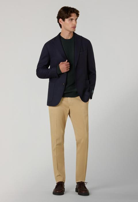 A man wearing an unbuttoned navy wool-twill jacket over a dark green crewneck knit, tan cotton trousers and dark brown leather moc-toe lace-up derbies on lug soles, standing on a plain grey ground.
