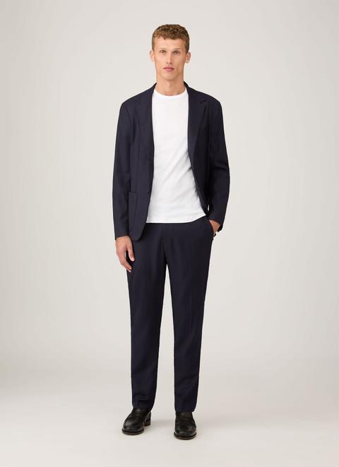 A man wearing a plain white crew-neck T-shirt under an open, unstructured navy jacket with patch pockets, matching navy trousers and black leather penny loafers, standing head to toe against a plain light-grey background.