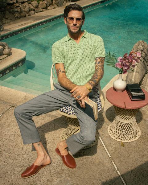 A man wearing a pistachio-green open-weave crochet knit polo, light-grey check trousers and tan leather backless slippers without socks, with tinted sunglasses and a gold watch, seated in a wicker chair beside a turquoise swimming pool.