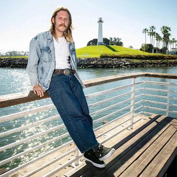 A man wearing a light stonewash denim jacket over a plain white tee, a brown western belt with a silver buckle, dark baggy straight-leg jeans and black suede Emerica low-top skate shoes, leaning on a harbour railing in sunshine with a lighthouse and palm trees behind him.