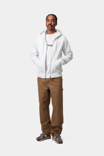A man wearing a light grey zip hoodie open over a white Carhartt logo tee, brown double-knee canvas work pants and grey-brown New Balance runners, standing head to toe on a plain light grey studio background.