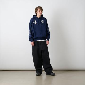 A man wearing a navy hoodie with a white number 4 and skull-crest graphic on the chest and spiderweb embroidery on the sleeves, a white T-shirt peeking out at the hem, very wide black jeans and black trainers, standing against a white studio wall.