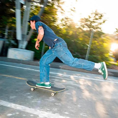 A man wearing a navy cap, a navy short-sleeve tee, baggy mid-wash straight-leg jeans, white socks and green suede low-top skate shoes, pushing a skateboard down a sunlit street.