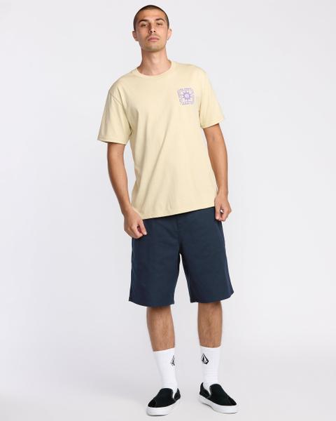 A man wearing a pale yellow t-shirt with a lilac Volcom chest graphic, navy knee-length chino shorts, white Volcom crew socks and black slip-on skate shoes with white soles, standing on a pale grey studio background.