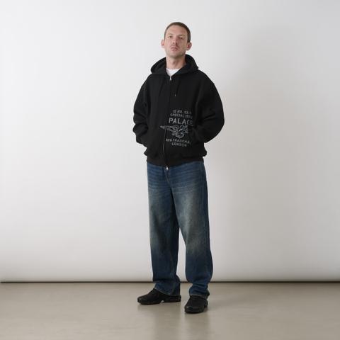 A man wearing an oversized black Palace zip hoodie with a grey eagle-and-logo print over a white T-shirt, baggy straight-leg faded indigo jeans and black Nike trainers, standing head to toe against a white studio wall.