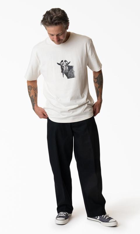 A man wearing a cream T-shirt with a black goat photo graphic, loose straight-leg black twill work pants and navy canvas low-top Converse sneakers, standing on a white studio background and looking down.