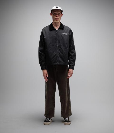 A man wearing a white six-panel cap, a boxy black zip work jacket with a corduroy collar over a white tee, loose dark brown corduroy trousers and black low-top skate shoes with white side stripes, standing on a grey studio background.
